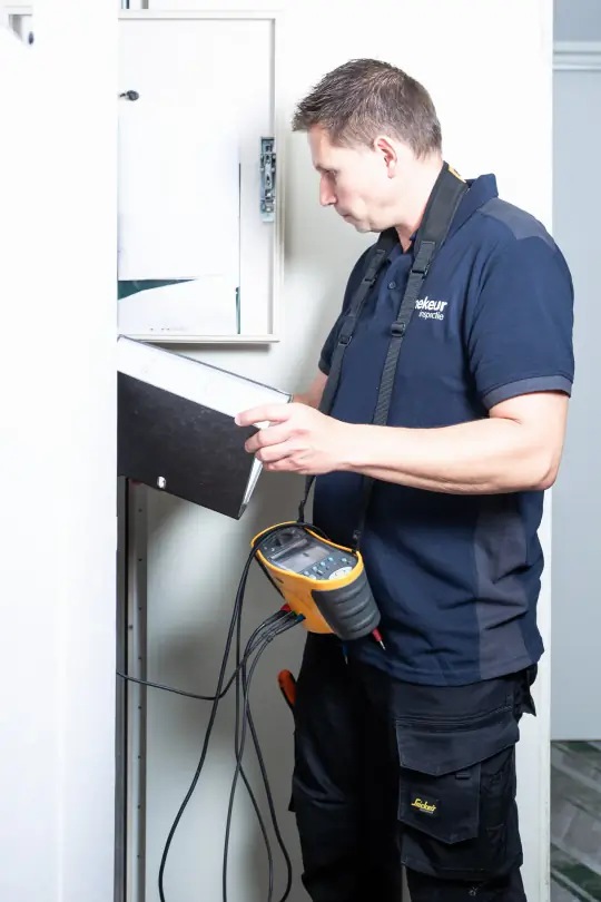 Inspecteur van Mekeur Inspectie leest het schema bij een groepenkast met een installatietester in de hand